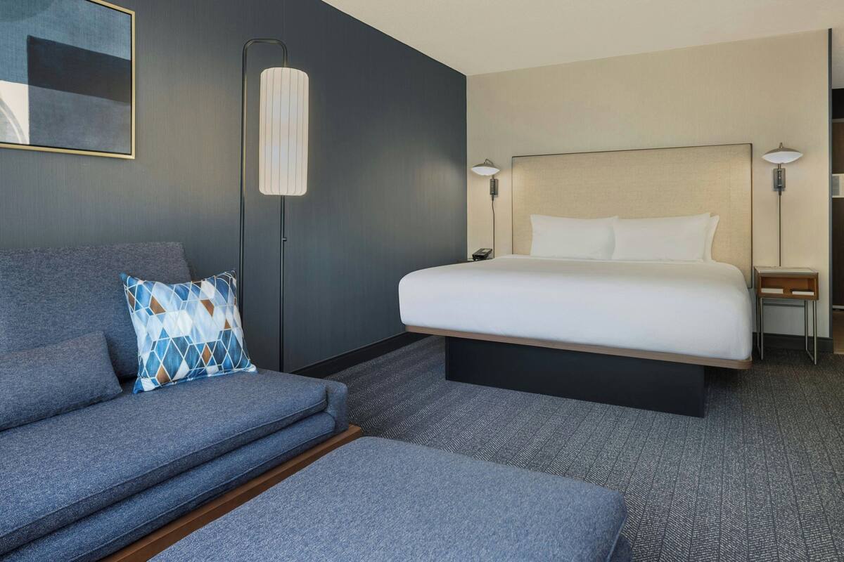 A modern hotel room with a large bed, a grey sofa, and a small table.