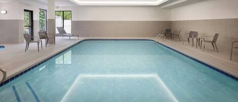 An indoor swimming pool with clear blue water, surrounded by lounge chairs and a tiled wall.