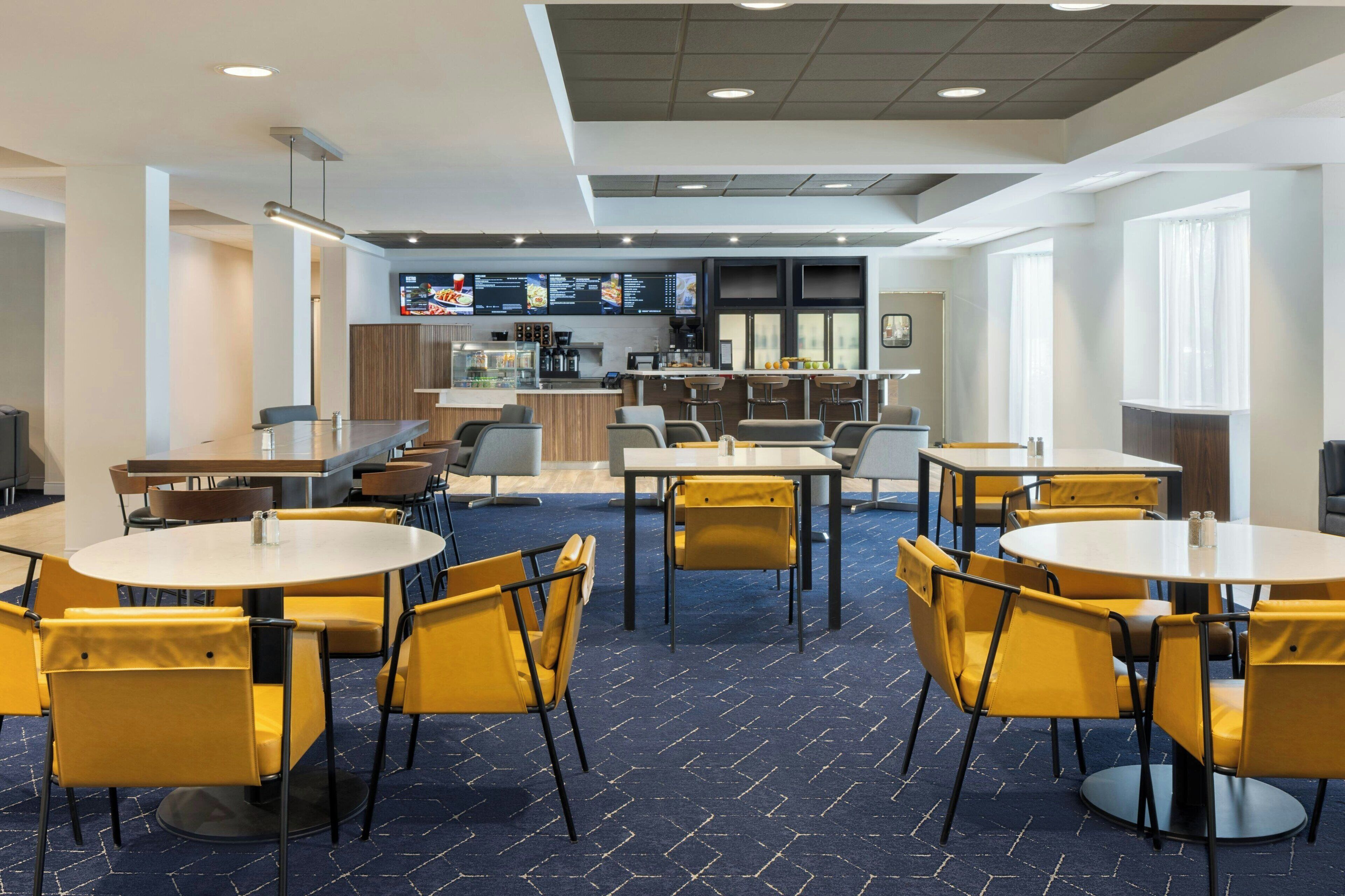 A modern dining area with round tables and yellow chairs, a bar area with stools, and a menu display.