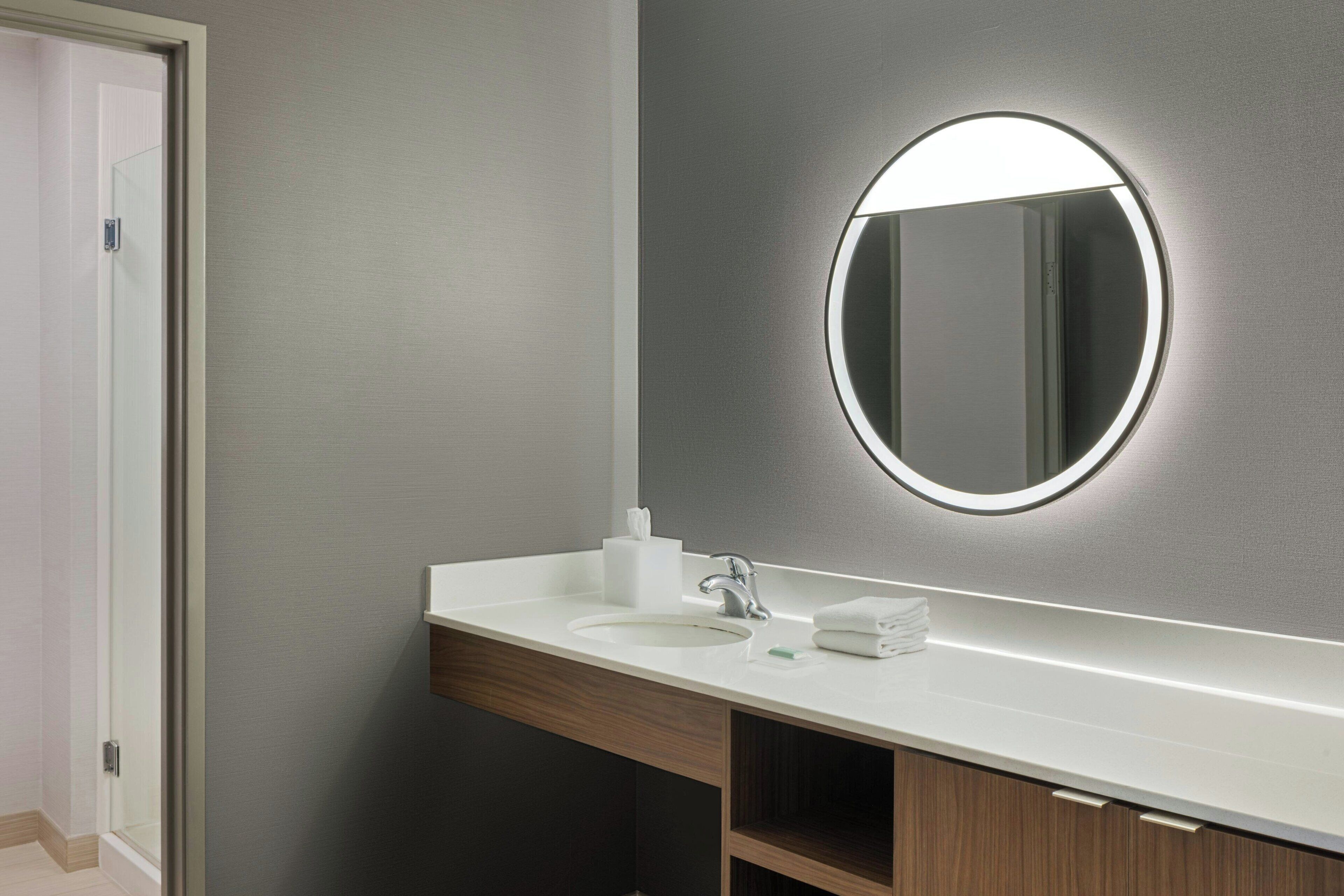 Un baño moderno con un lavamanos blanco, un espejo redondo con luz y un mueble de madera debajo del lavamanos.