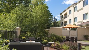 An outdoor patio with a sofa, table, and umbrella, surrounded by greenery and a building in the background. - Courtyard by Marriott Traverse City (Traverse City)