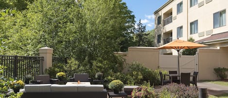 Une terrasse extérieure avec un canapé, une table et un parasol, entourée de verdure et avec un bâtiment en arrière-plan.