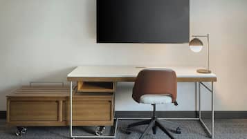 A modern office desk with a flat-screen TV, a brown office chair, a desk lamp, and a small wooden storage unit with wheels.