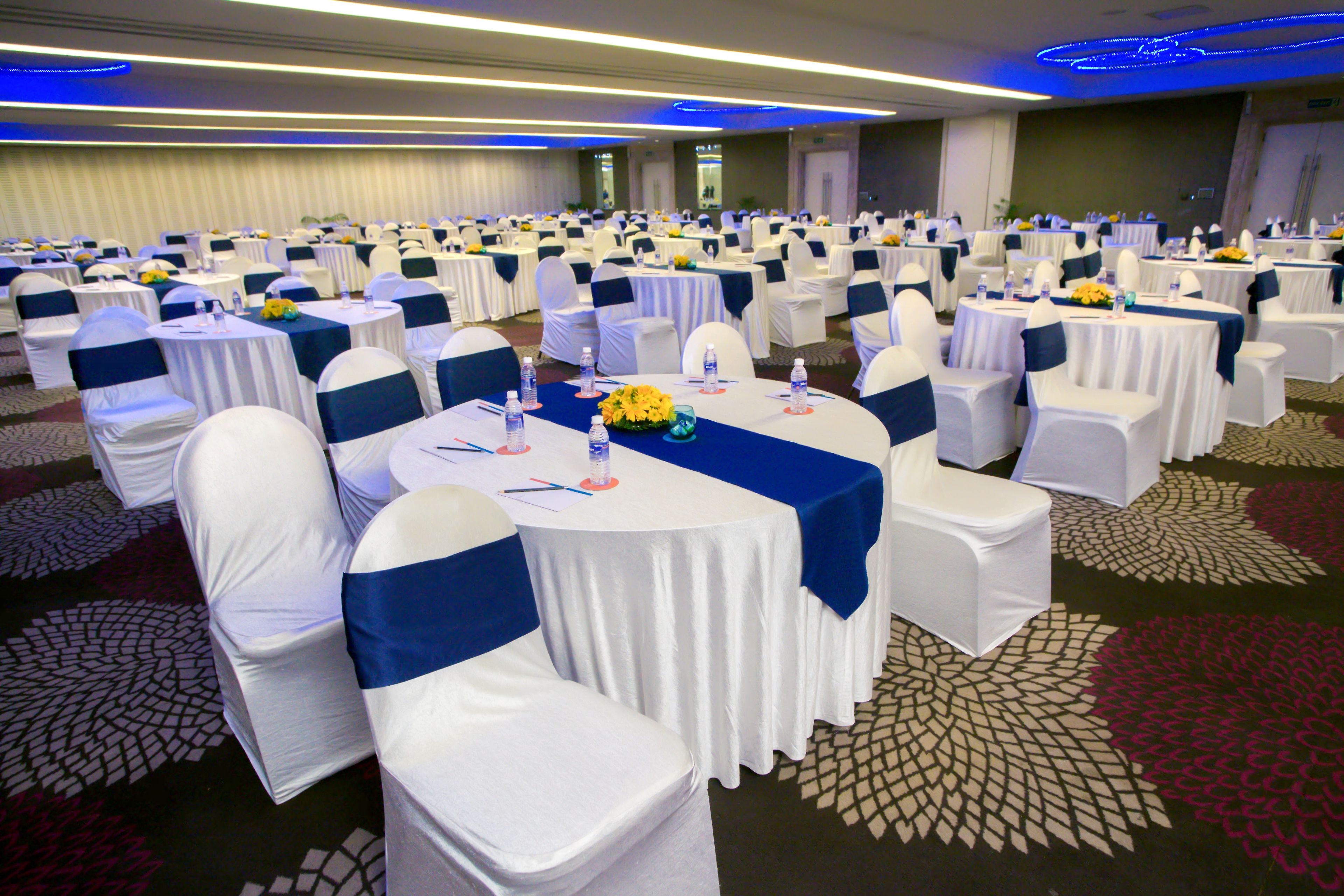 A banquet hall with round tables, white chairs, and blue sashes. Tables are set with water bottles and floral centerpieces.