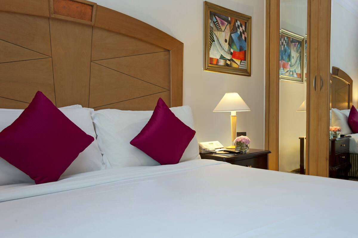 Habitación de hotel con una cama grande, dos almohadas, una lámpara de noche, un teléfono y un cuadro en la pared.