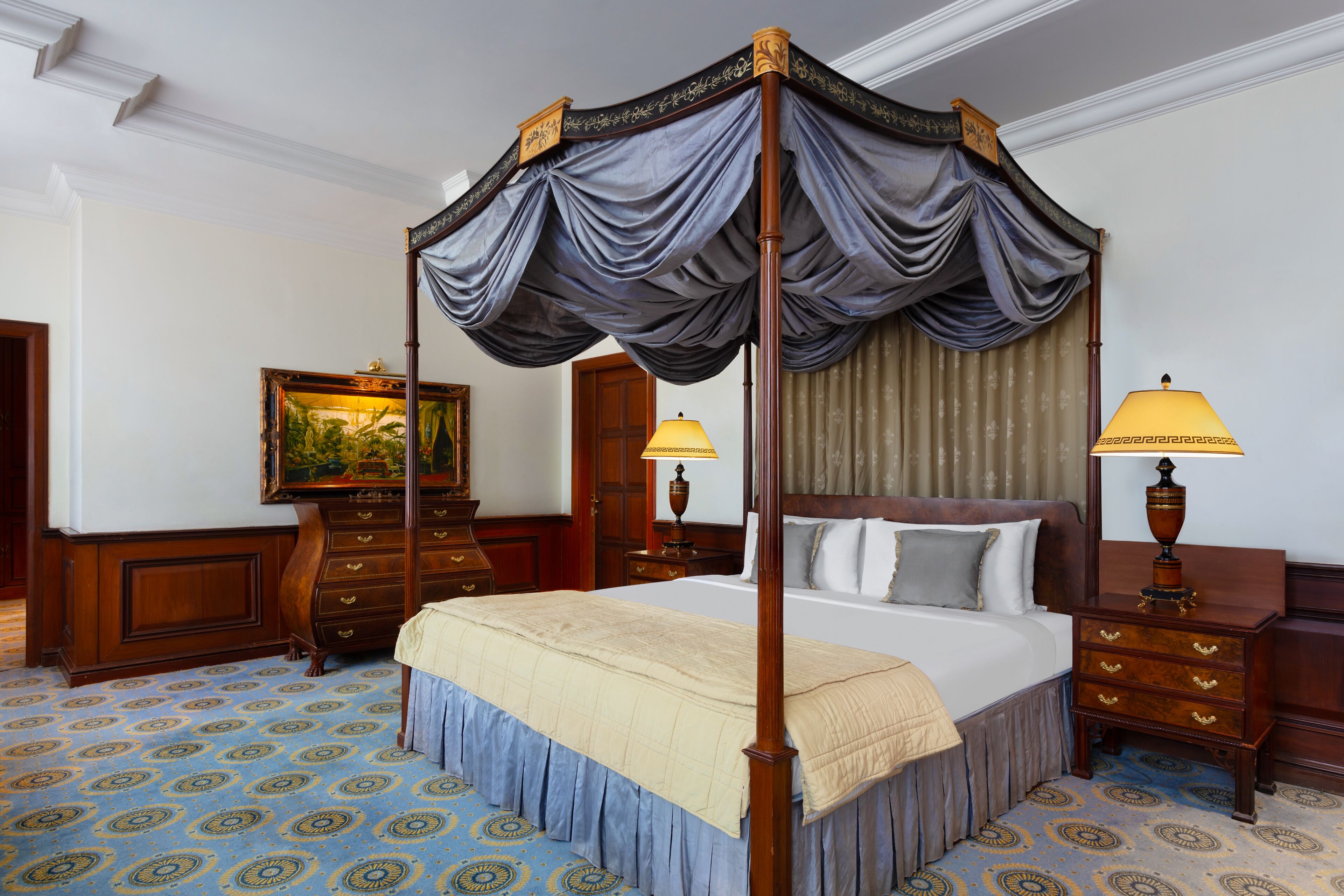 Una cama con dosel y cuatro columnas, cubierta con una colcha color beige, dos lámparas de noche, una mesita de madera y una alfombra estampada.