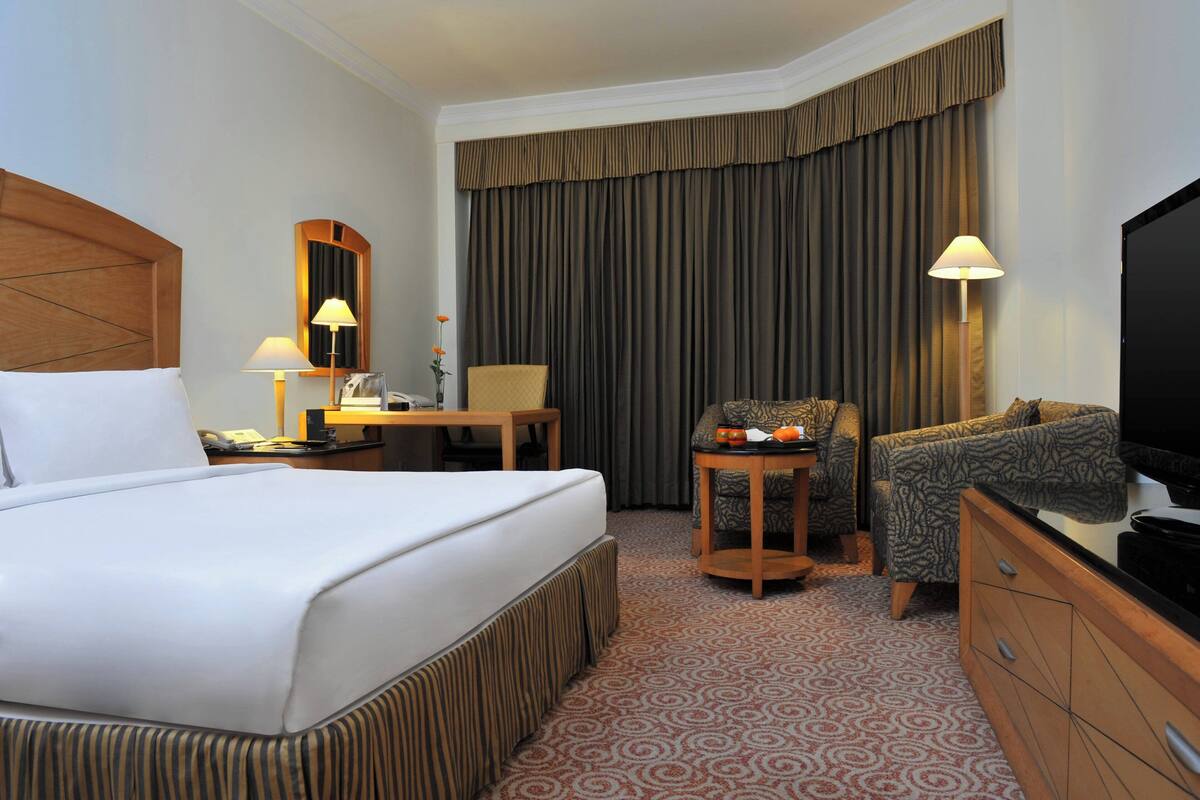 Habitación de hotel con una cama grande, un escritorio, una silla, un televisor y un ventanal con cortinas.