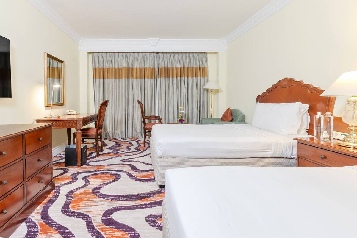 Habitación de hotel con dos camas, un escritorio, una silla, un aparador y un ventanal con cortinas.