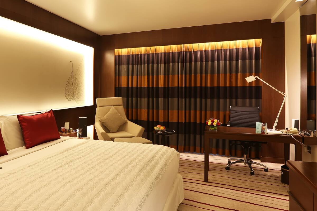 Habitación de hotel con una cama grande, un escritorio con silla, una lámpara y vistas al exterior a través de cortinas.