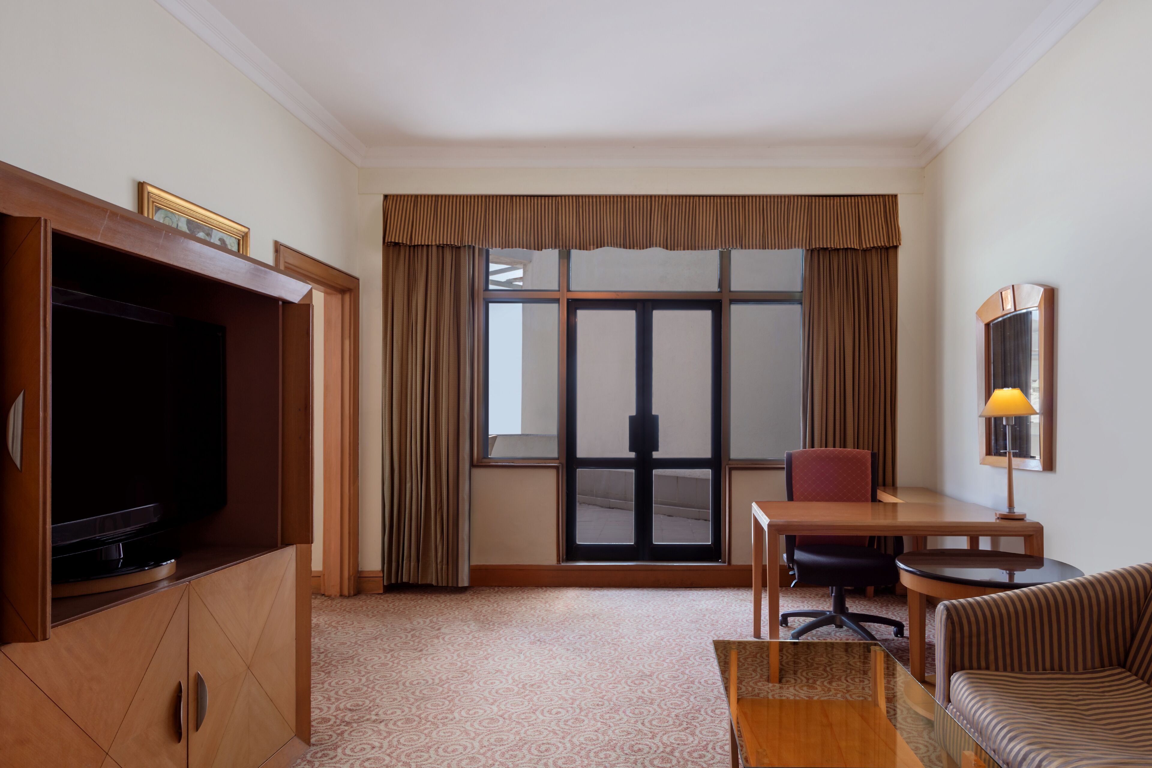 Habitación de hotel con televisión, escritorio, silla, sofá y un amplio ventanal con cortinas.