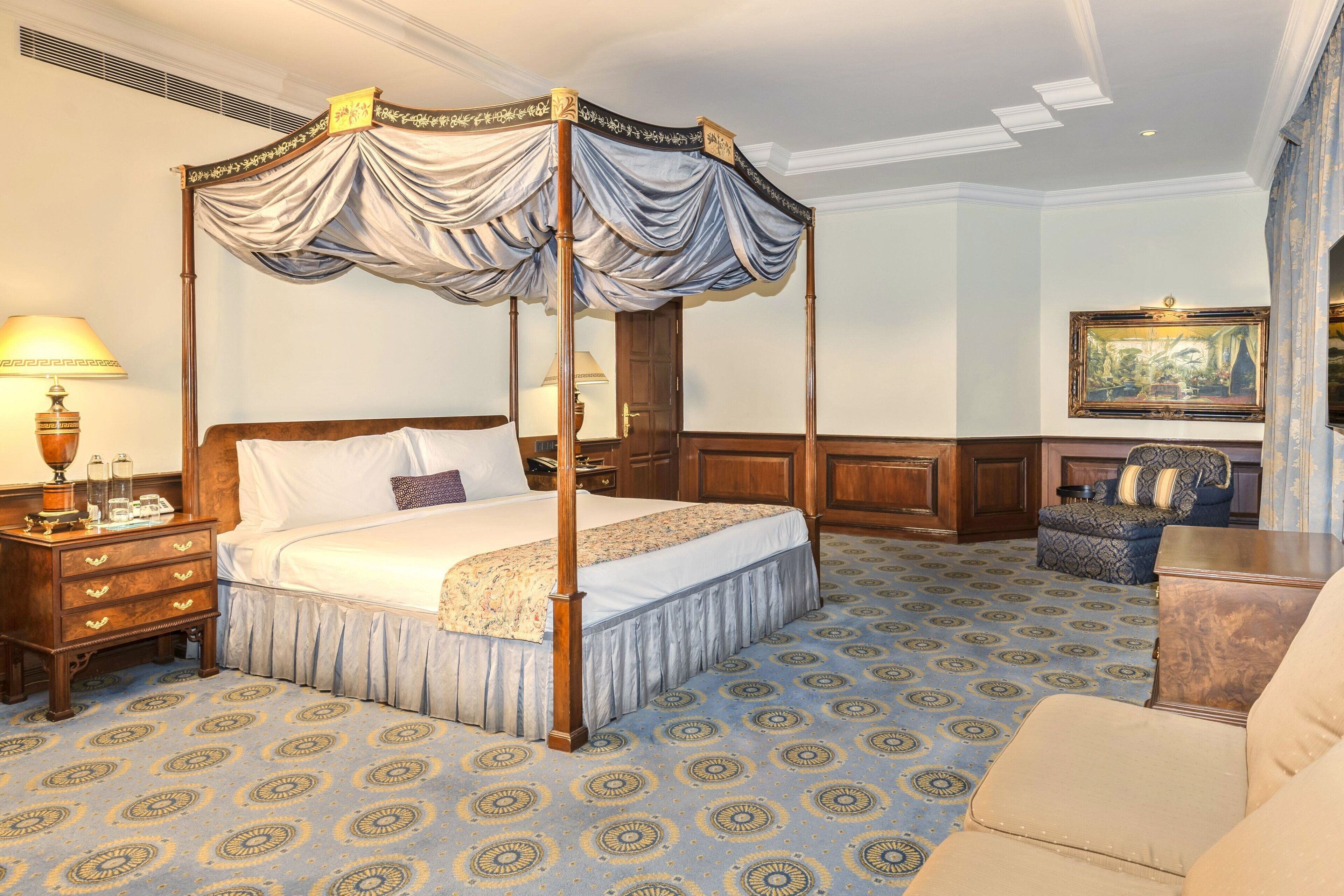 A four-poster canopy bed with a wooden frame and draped fabric, a nightstand with a lamp, a patterned carpet, and a seating area with a sofa and armchair.