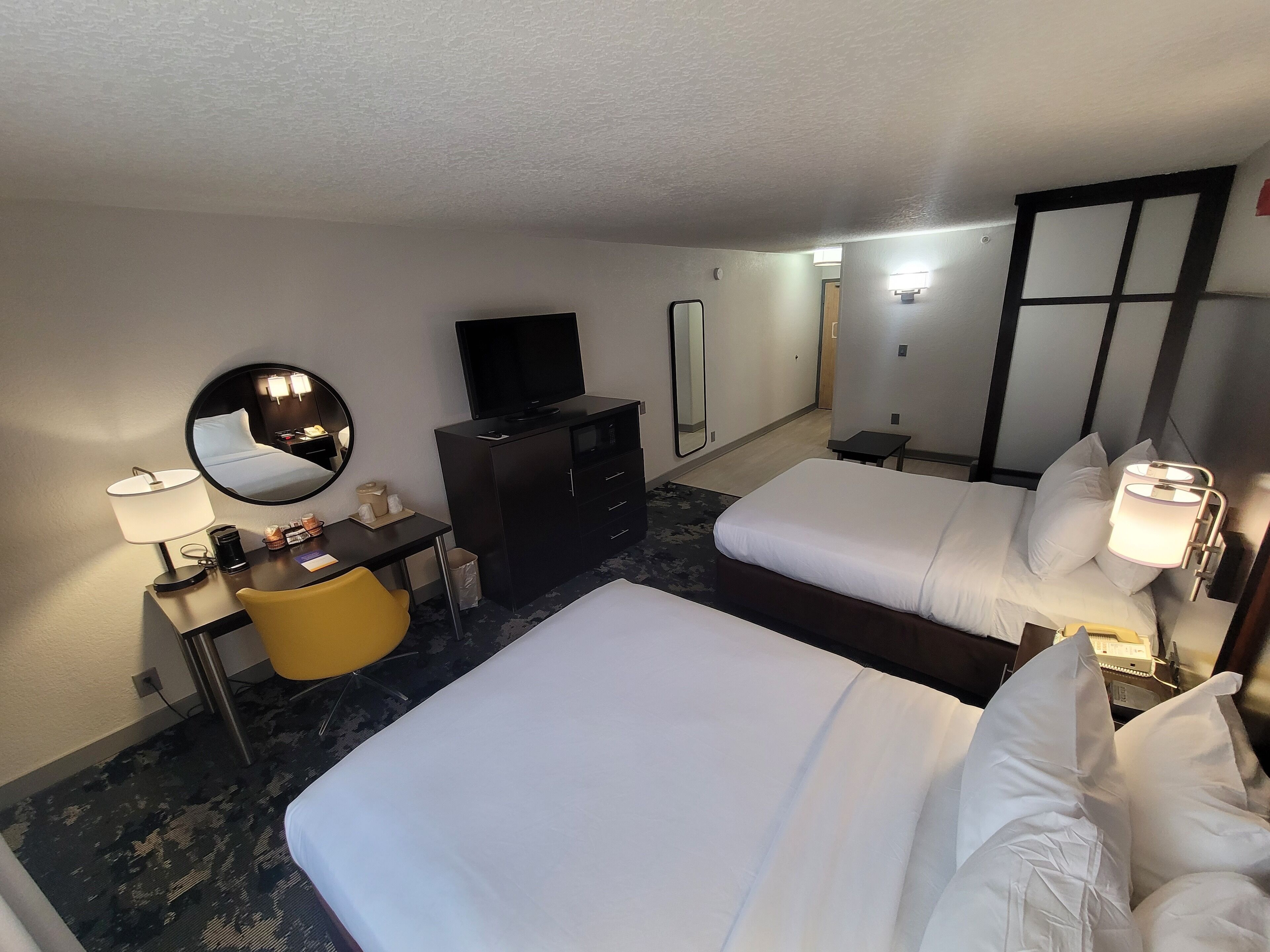 Quarto de hotel com duas camas, uma escrivaninha com luminária, televisão, espelho e uma mesinha.