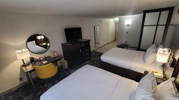 Quarto de hotel com duas camas, uma escrivaninha com luminária, televisão, espelho e uma mesinha.