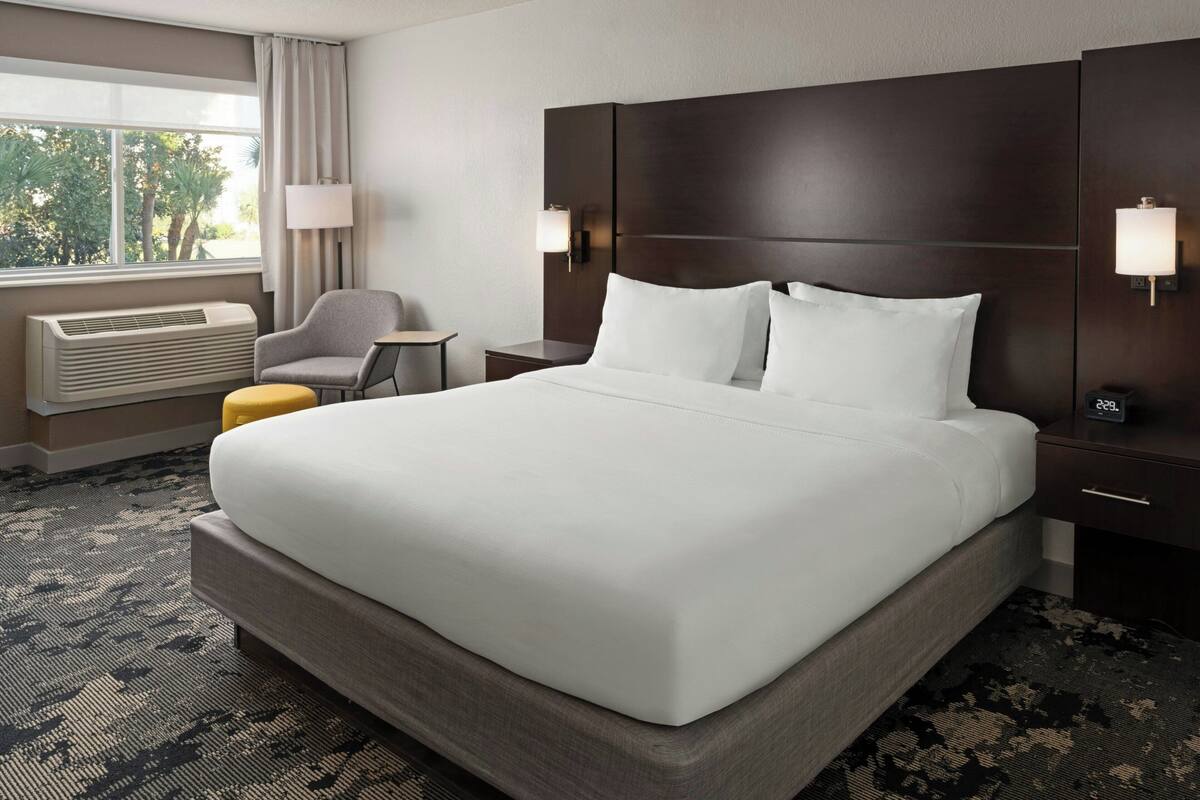 Hotelkamer met een groot bed, een bureau, een stoel, een lamp en uitzicht op bomen door het raam.