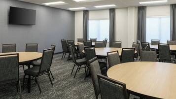 A conference room with multiple tables and chairs, a flat-screen TV mounted on the wall, and large windows with curtains.