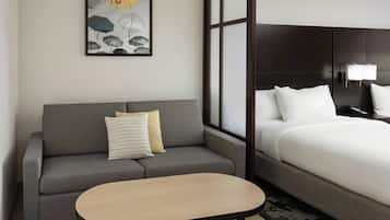 A hotel room with a grey sofa, a small round table, a bed with white bedding, a dark headboard, and a wall-mounted lamp.