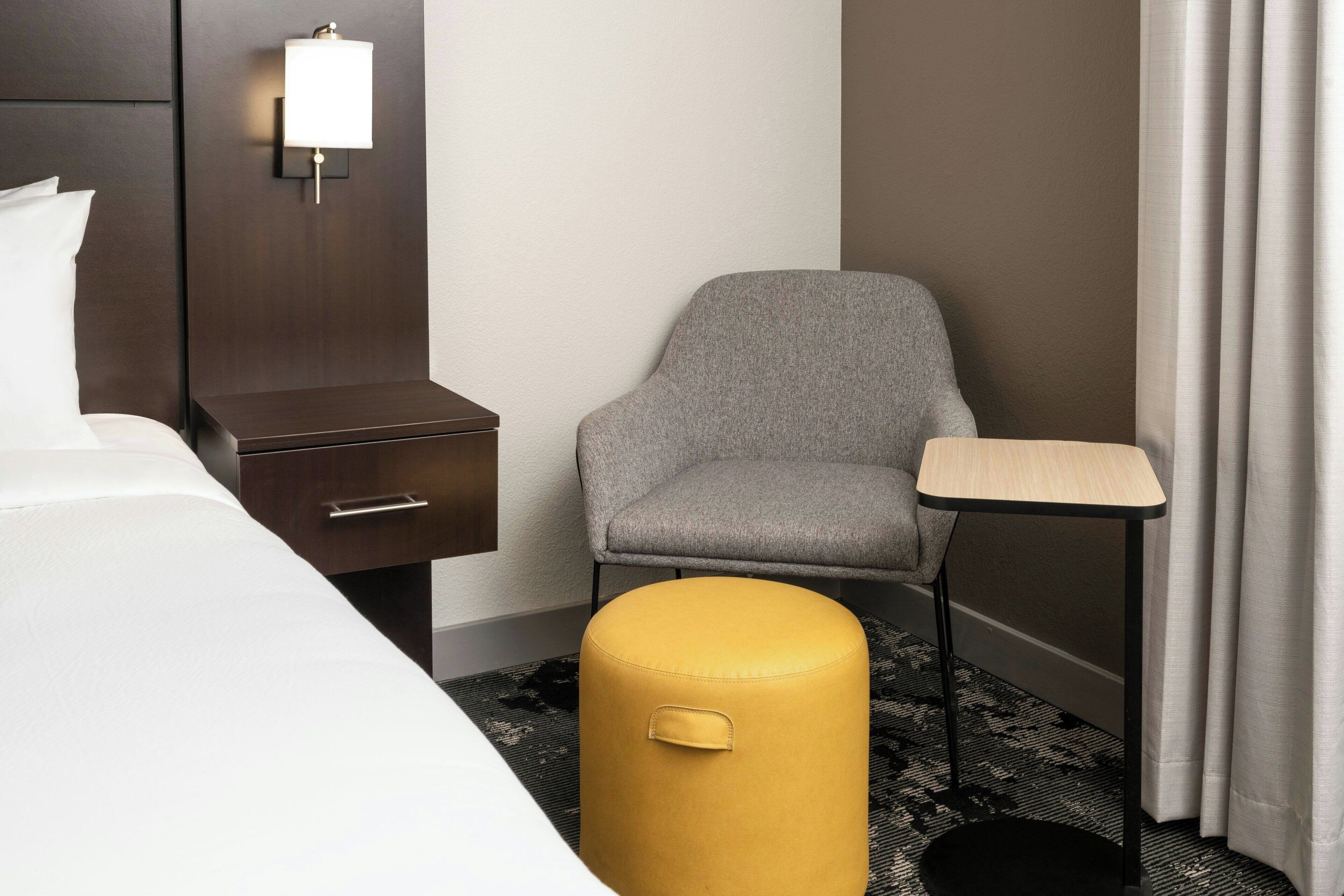 A hotel room with a bed, a nightstand, a chair, a small table, and a yellow ottoman.