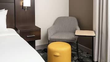 Une chambre d’hôtel avec un lit, une table de chevet, une chaise, une petite table et un pouf jaune.