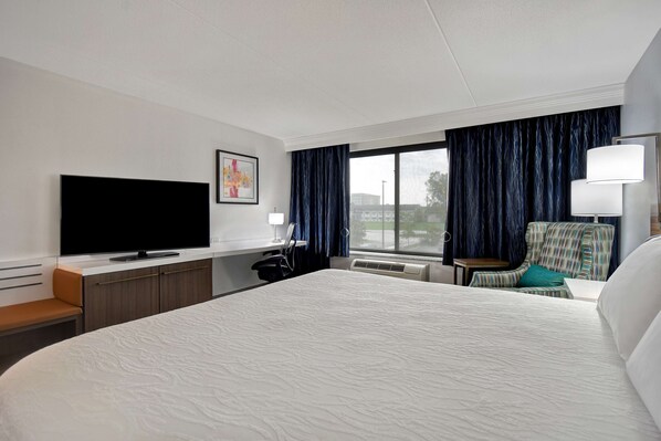 A hotel room with a large bed, a flat-screen TV, a desk with a chair, and a window with curtains.
