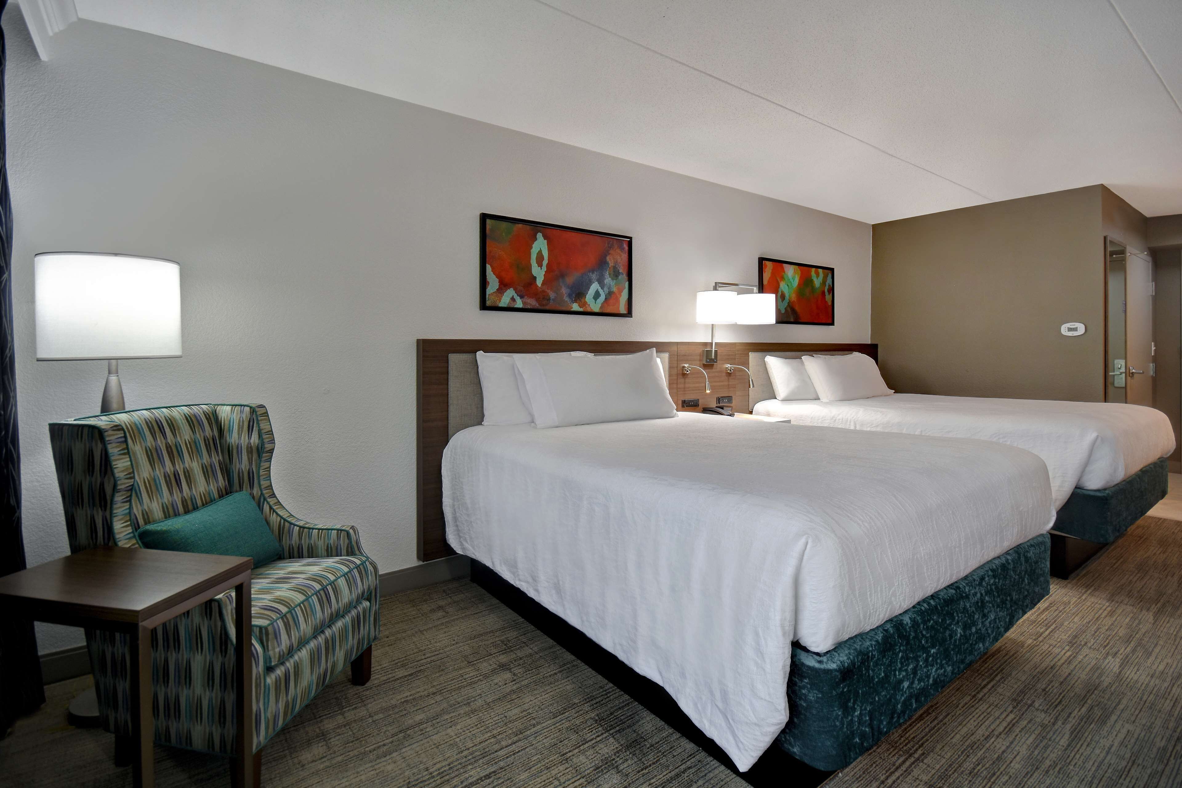 A hotel room with two beds, a chair, a table, and two wall-mounted lamps.