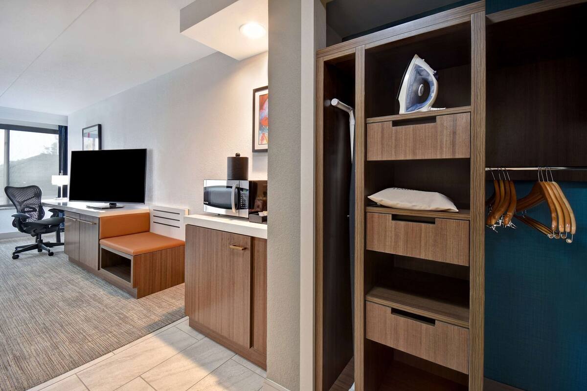 A modern hotel room with a desk, a flat-screen TV, a microwave, a seating area, and a wardrobe with hanging clothes.