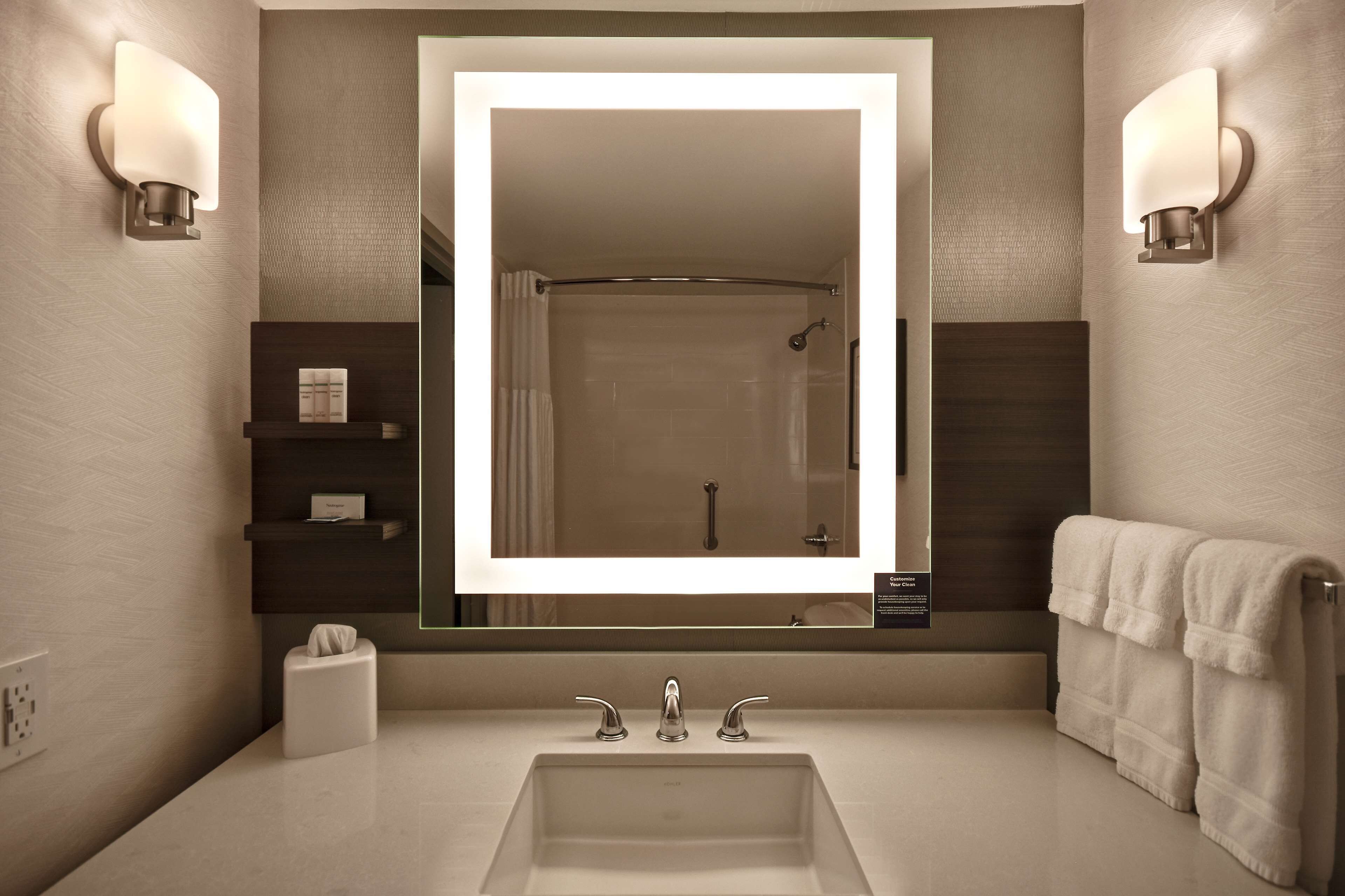 A modern bathroom with a large mirror, two wall-mounted lights, a towel rack, and a shelf with toiletries.