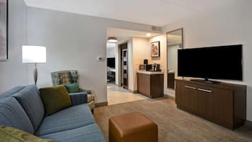 A modern hotel room with a sofa, ottoman, flat-screen TV, and a kitchenette area.