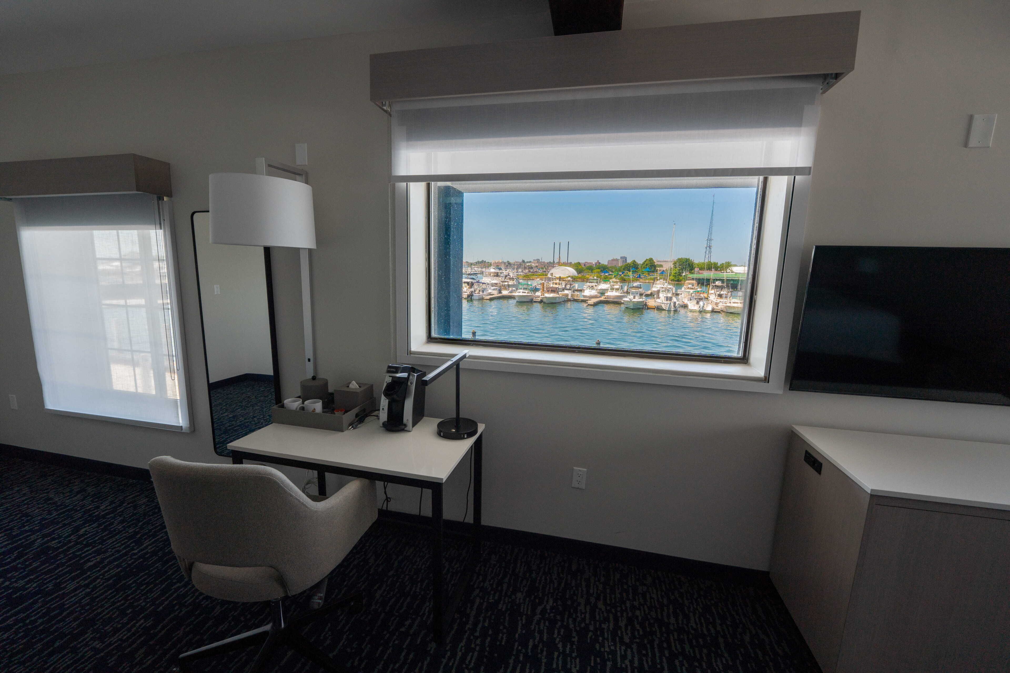 A hotel room with a desk, chair, TV, and a large window with a marina view.
