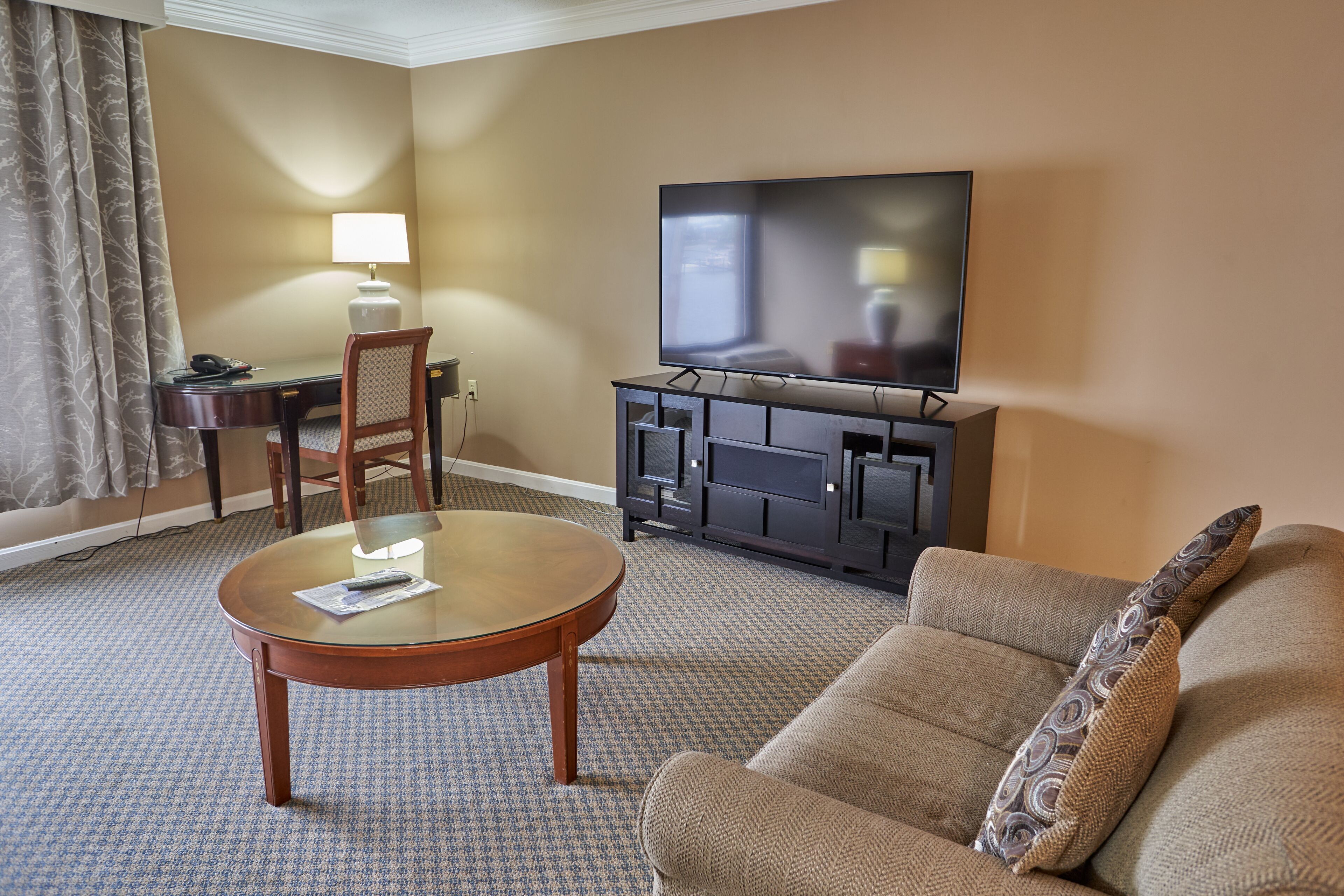 A hotel room with a flat-screen TV, a round glass table, a sofa, a desk with a lamp, and a chair.