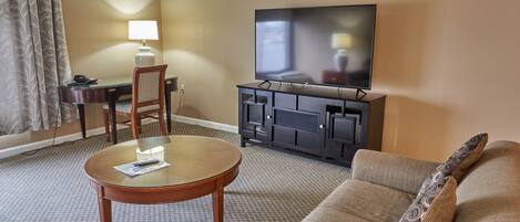 A hotel room with a flat-screen TV, a round glass table, a sofa, a desk with a lamp, and a chair.