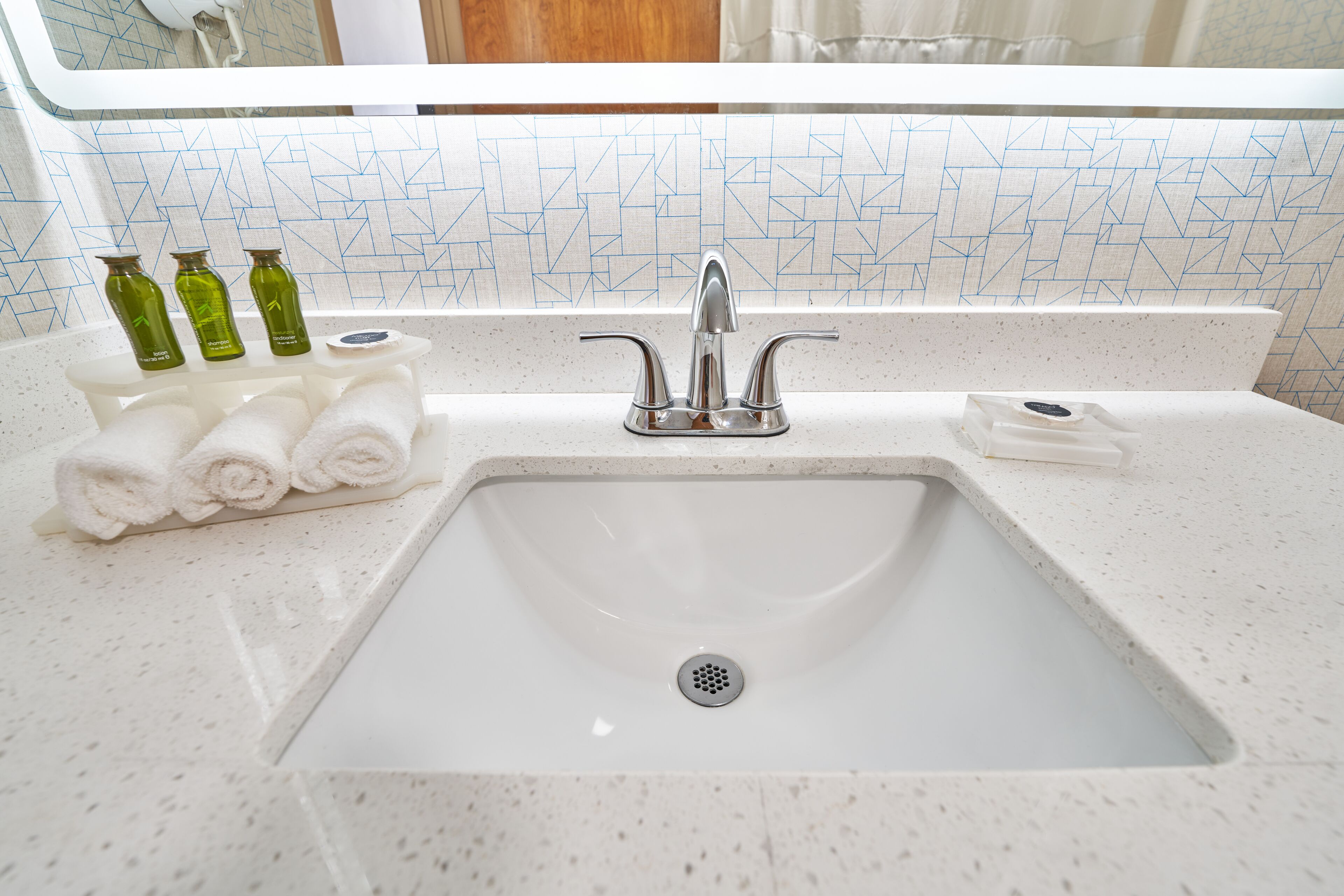 A bathroom sink with a white countertop, a mirror above it, and a tiled backsplash. There are three green bottles and several rolled towels on the counter.
