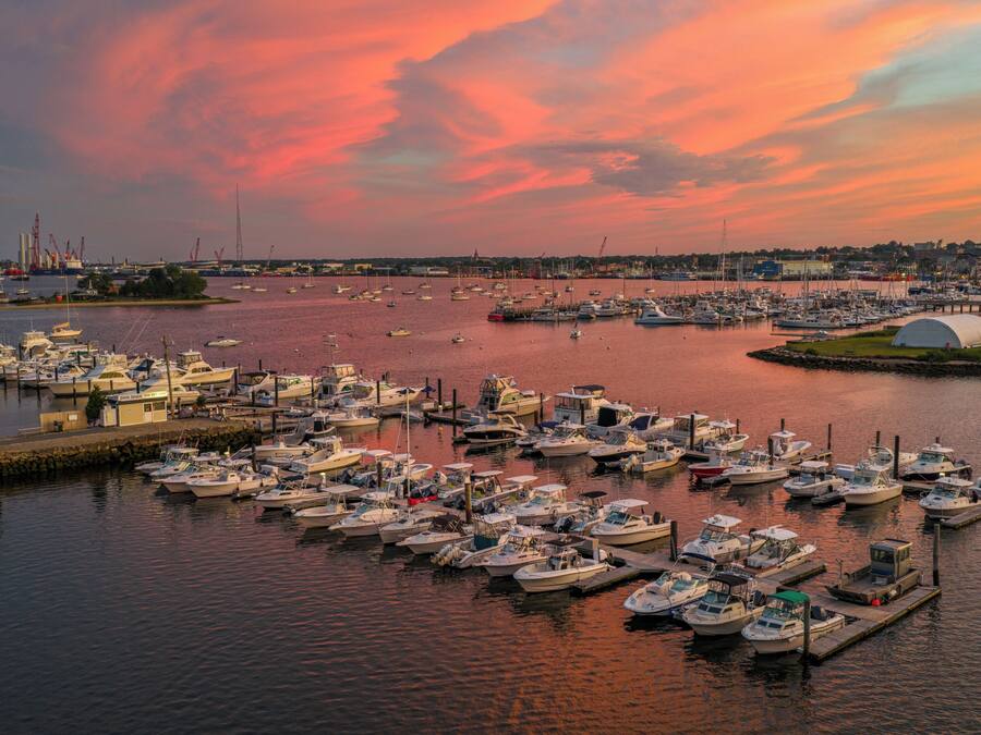 A marina with numerous boats docked, a distant cityscape, and a sunset sky.