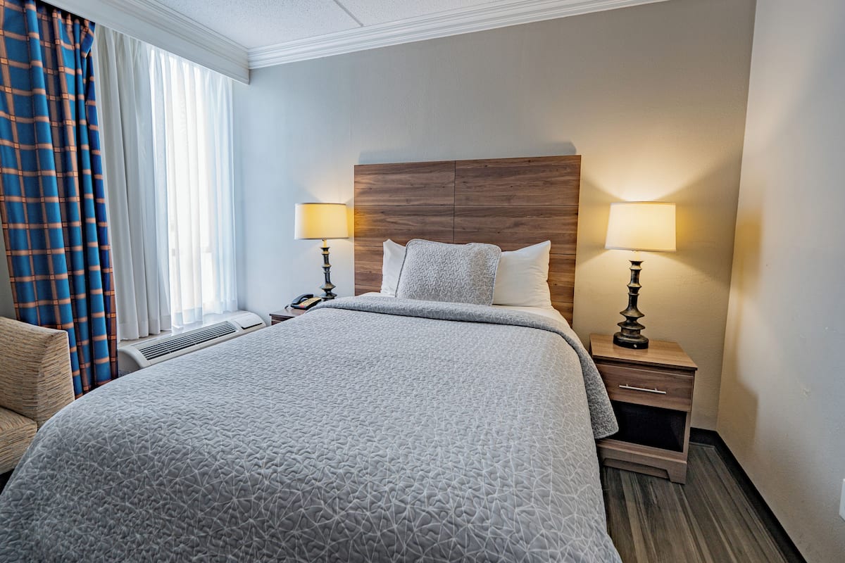 A hotel room with a large bed, two bedside tables with lamps, a wooden headboard, a chair, and a window with curtains.