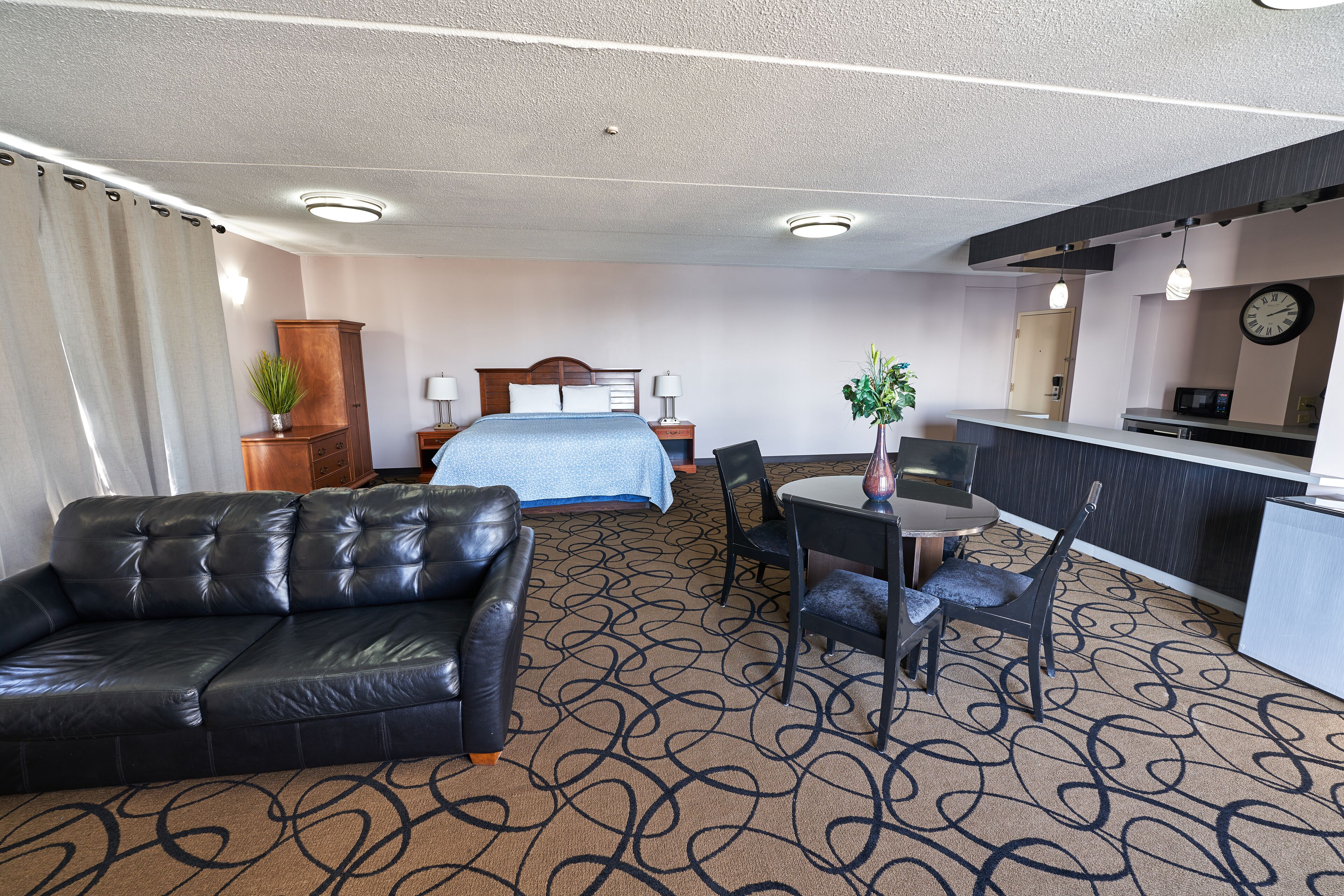 Habitación de hotel con cama, sofá de cuero, mesa de comedor con sillas, una pequeña cocina y mostrador de recepción.