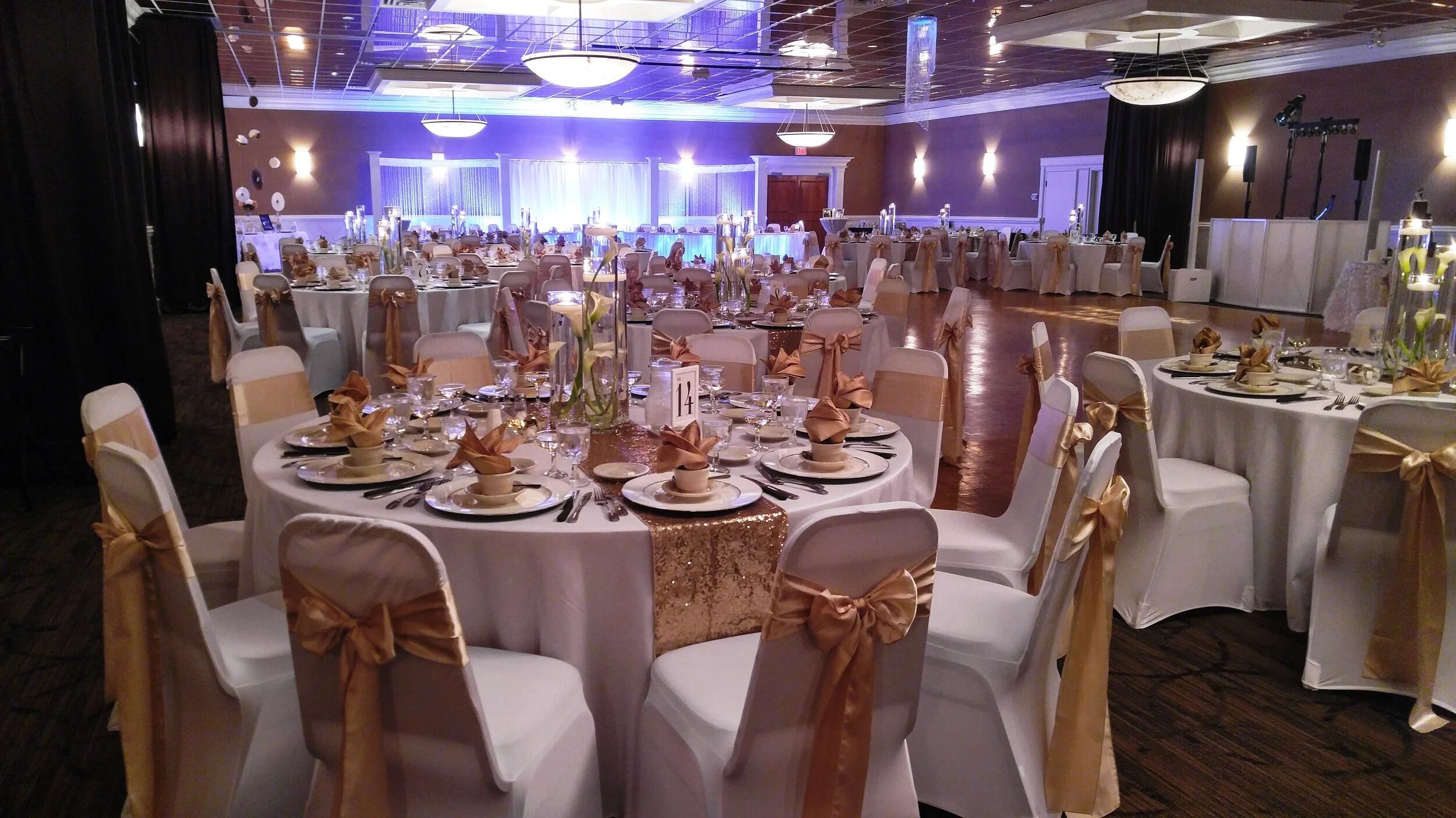 A banquet hall with round tables, chairs with gold sashes, and a stage in the background.