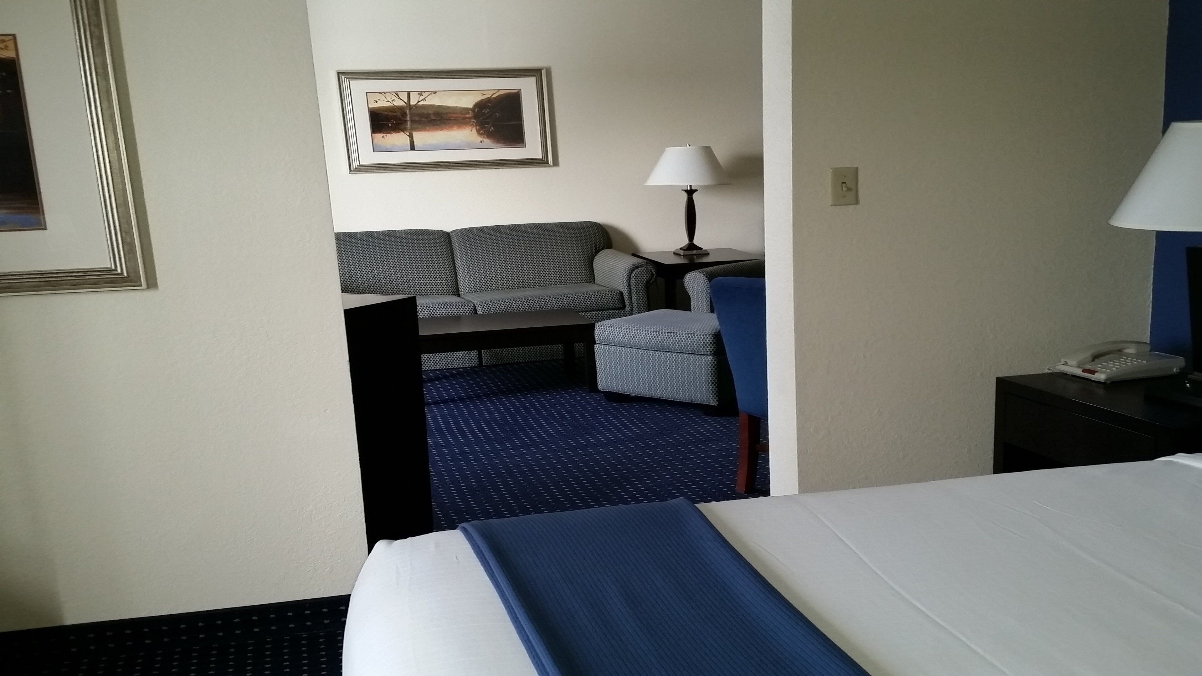 Ett hotellrum med en säng, ett skrivbord, en stol, en lampa och en inramad tavla på väggen.