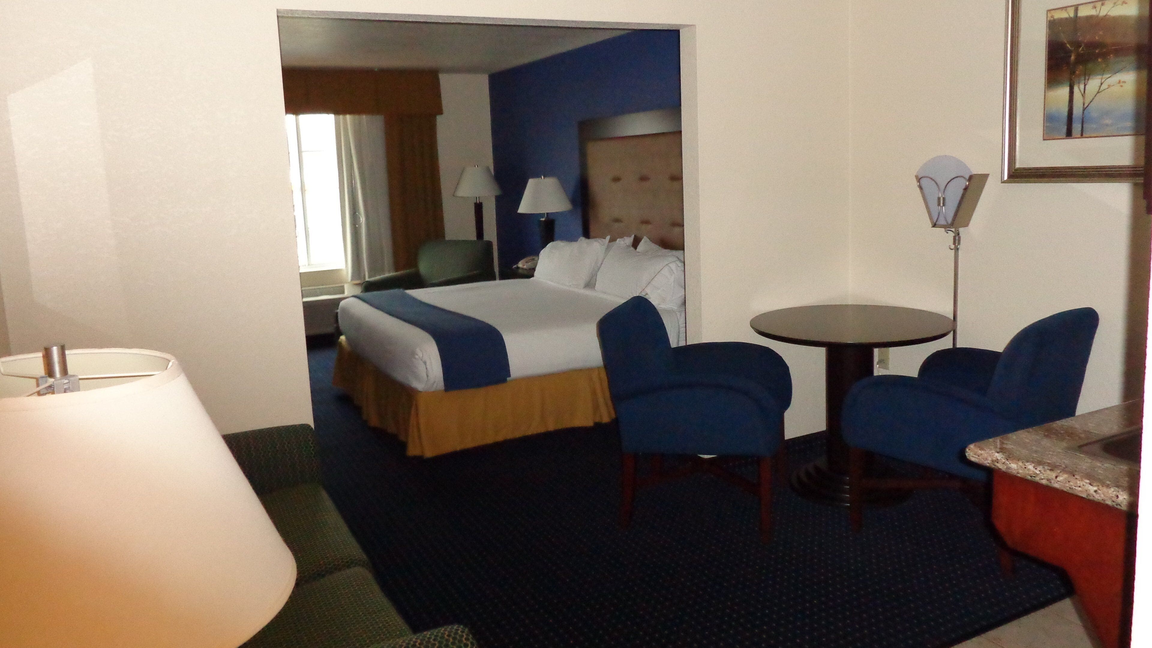 A hotel room with a bed, two chairs, a small table, a lamp, and a painting on the wall.