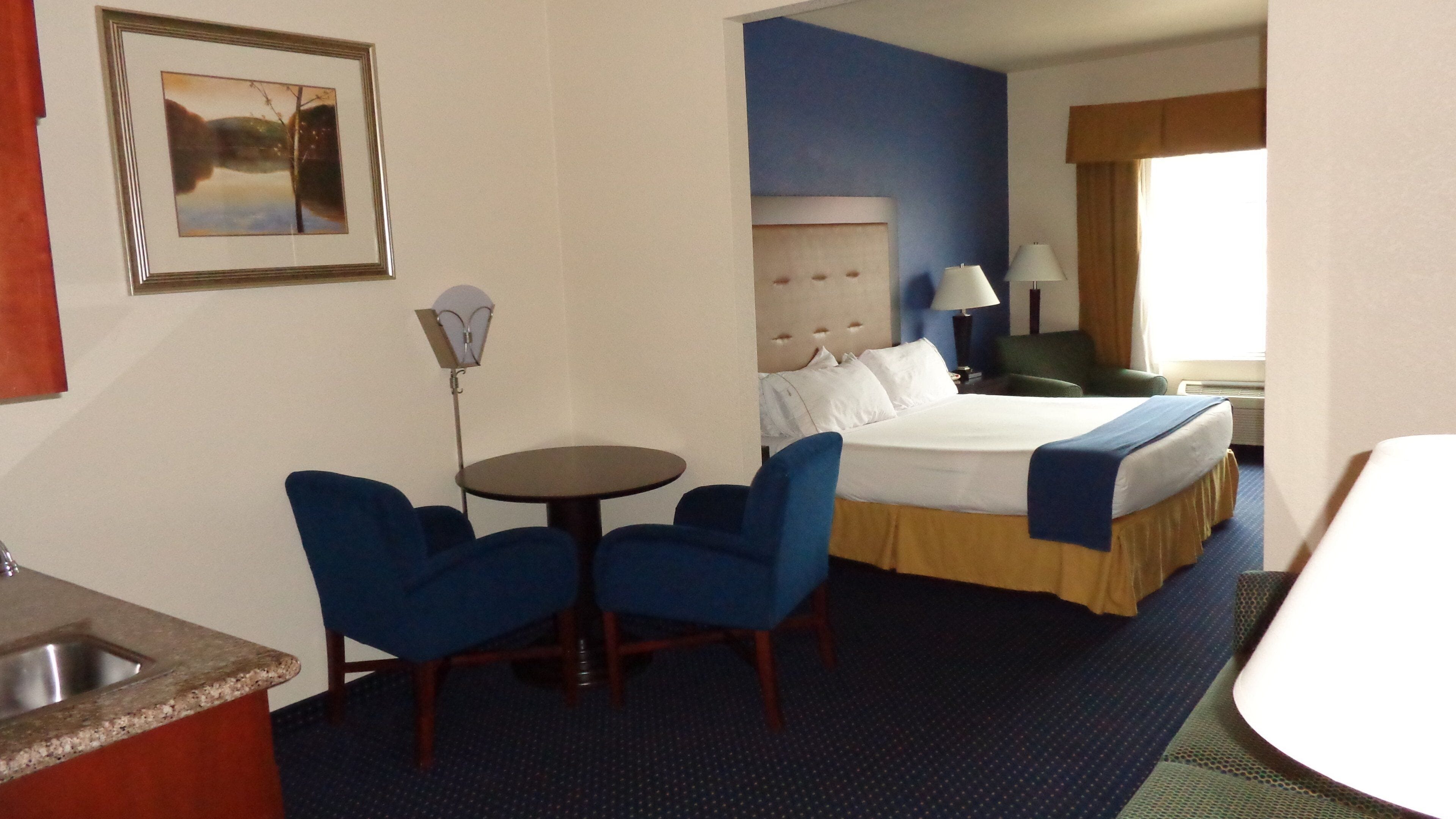 A hotel room with a bed, a desk, two chairs, a small table, a lamp, and a picture on the wall.