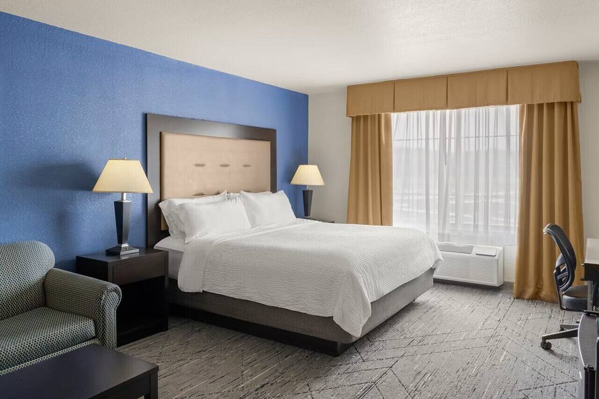 A hotel room with a bed, two bedside tables with lamps, a chair, a desk, and a large window with curtains.
