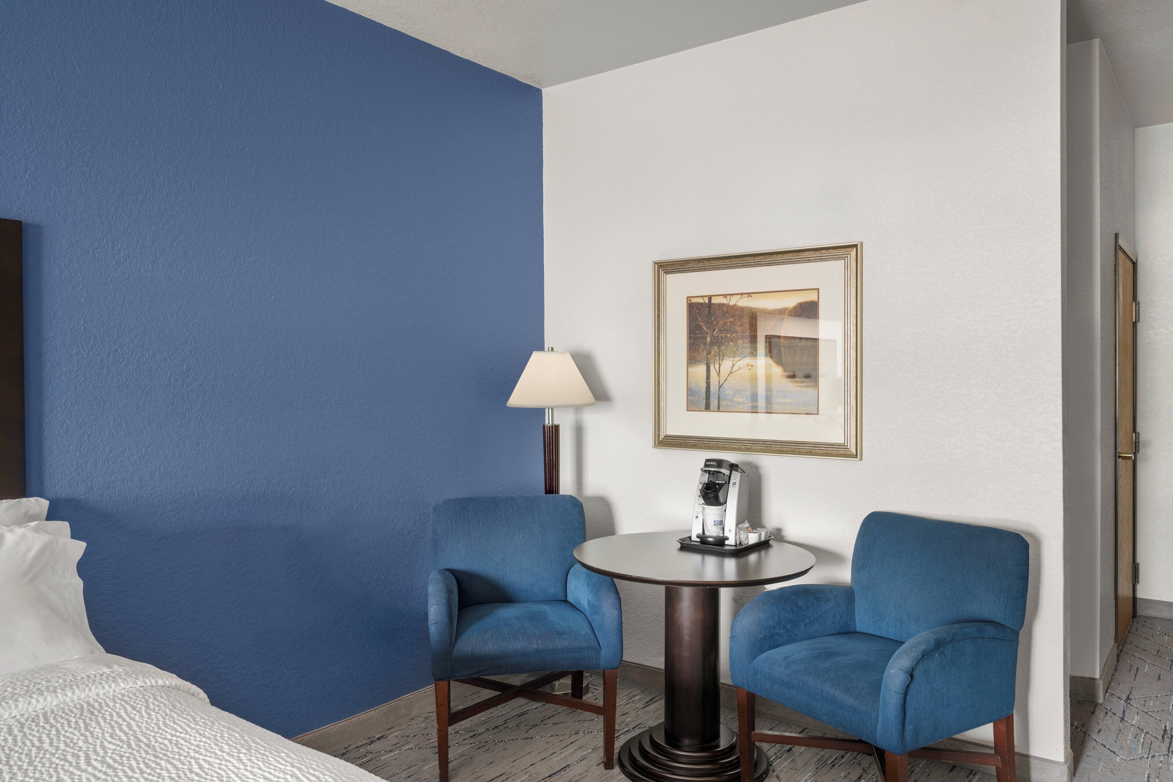 A hotel room with a blue accent wall, two blue armchairs, a round table with a coffee maker, a framed picture on the wall, and a bed with white bedding.