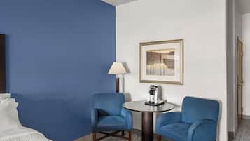 Habitación de hotel con una pared azul decorativa, dos sillones azules, una mesa redonda con cafetera, un cuadro enmarcado en la pared y una cama con ropa de cama blanca.