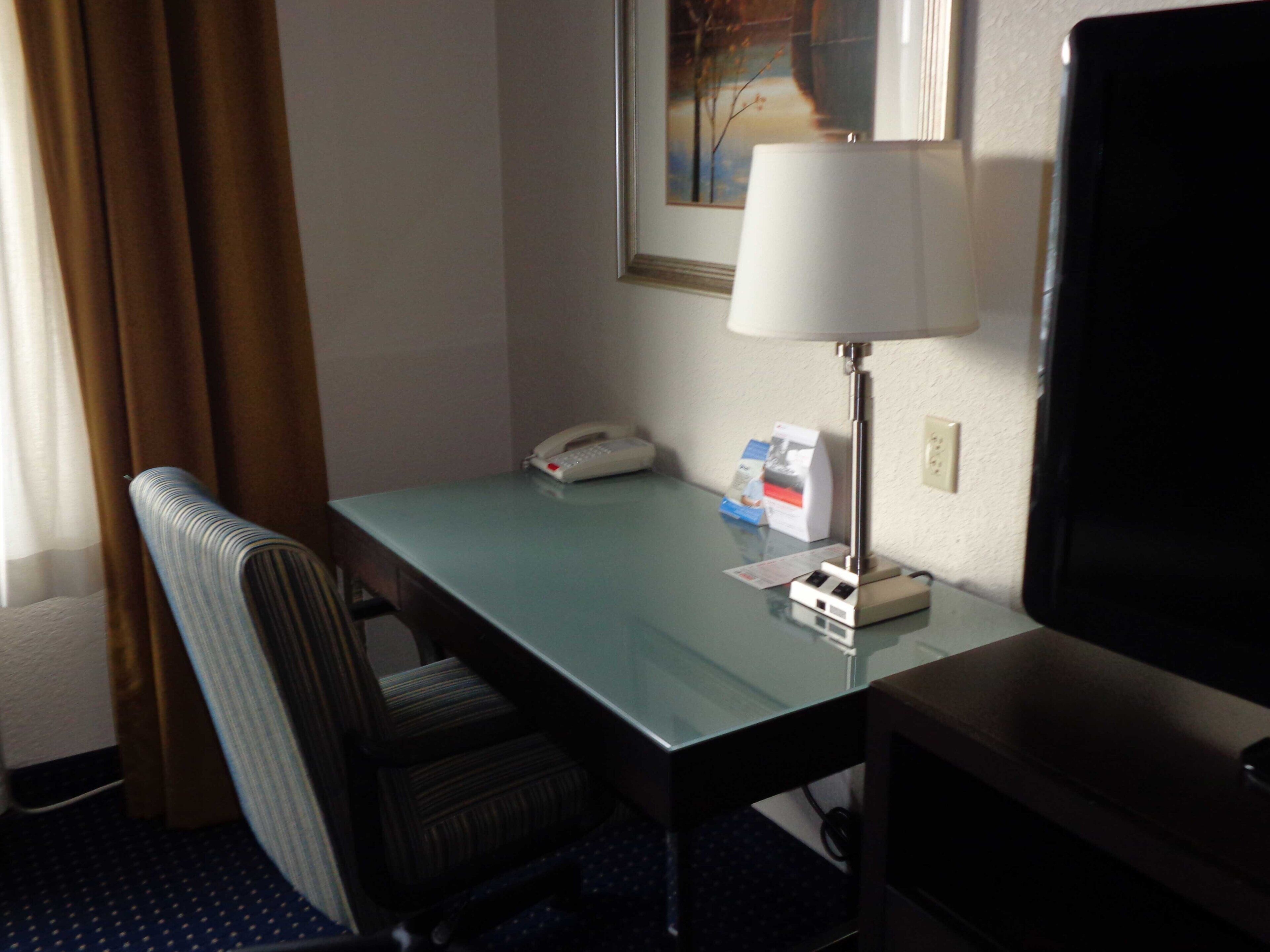 A hotel room with a glass desk, a telephone, a lamp, a television, and a chair.
