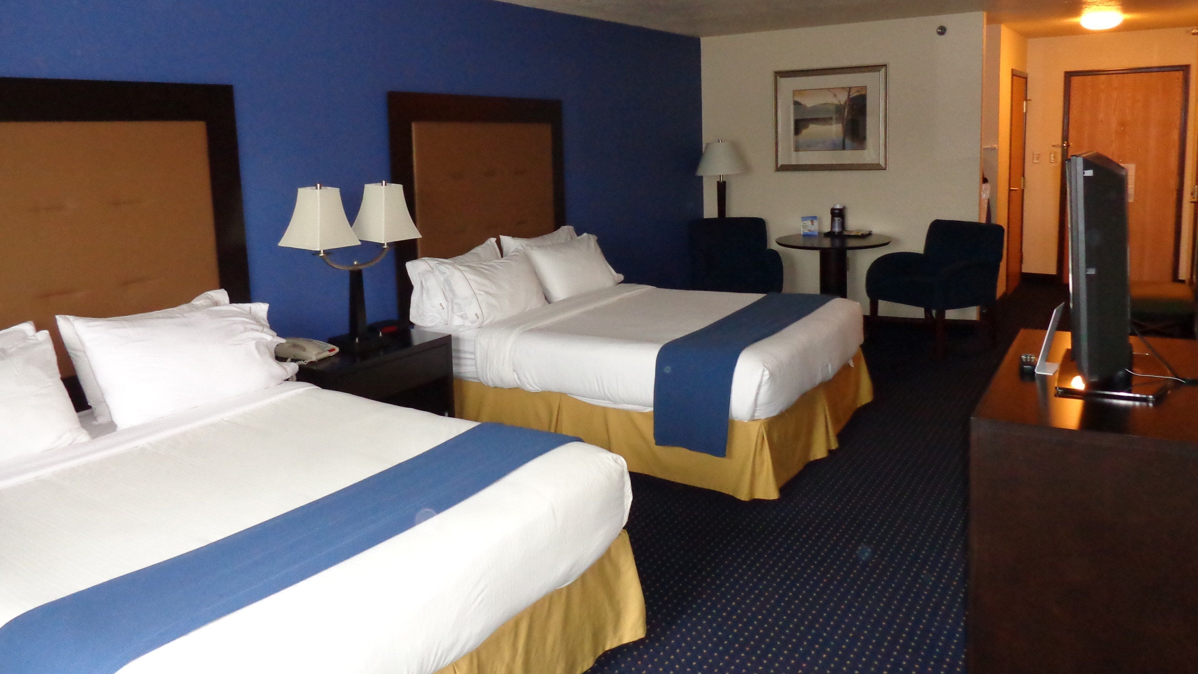 A hotel room with two beds, a desk with a computer, a chair, and a small table with a lamp.