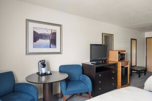 A hotel room with a bed, a desk with a coffee maker, two blue chairs, a television, a dresser, and a framed picture on the wall.