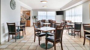 A dining area with multiple tables and chairs, a clock, a television, and a kitchen area in the background. - Quality Inn & Suites Manistique (Manistique)