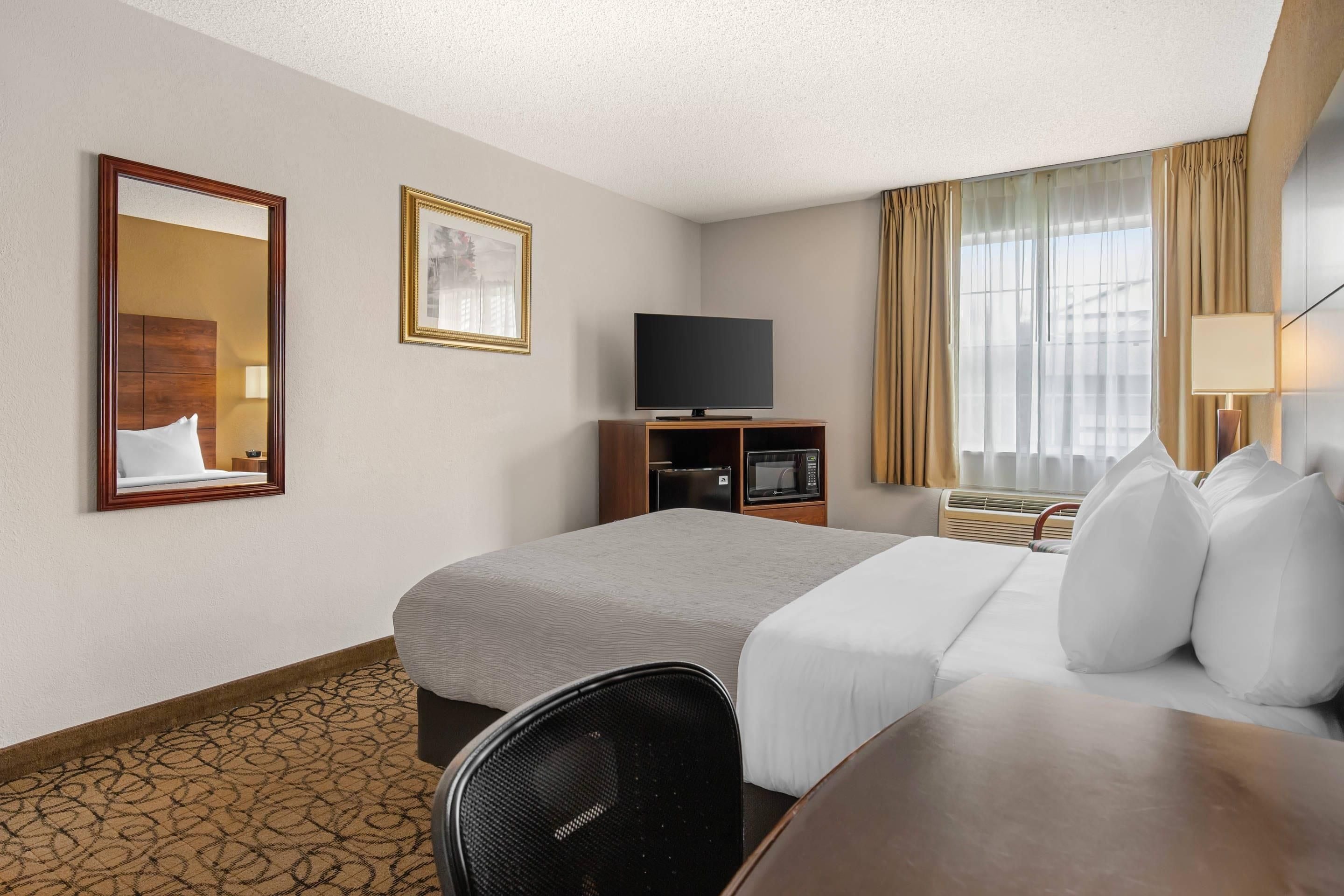 A hotel room with a bed, a TV, a desk, a chair, a mirror, and a window with curtains.