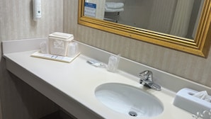 A hotel bathroom with a white sink, a mirror with a gold frame, a towel dispenser, and a soap dispenser.