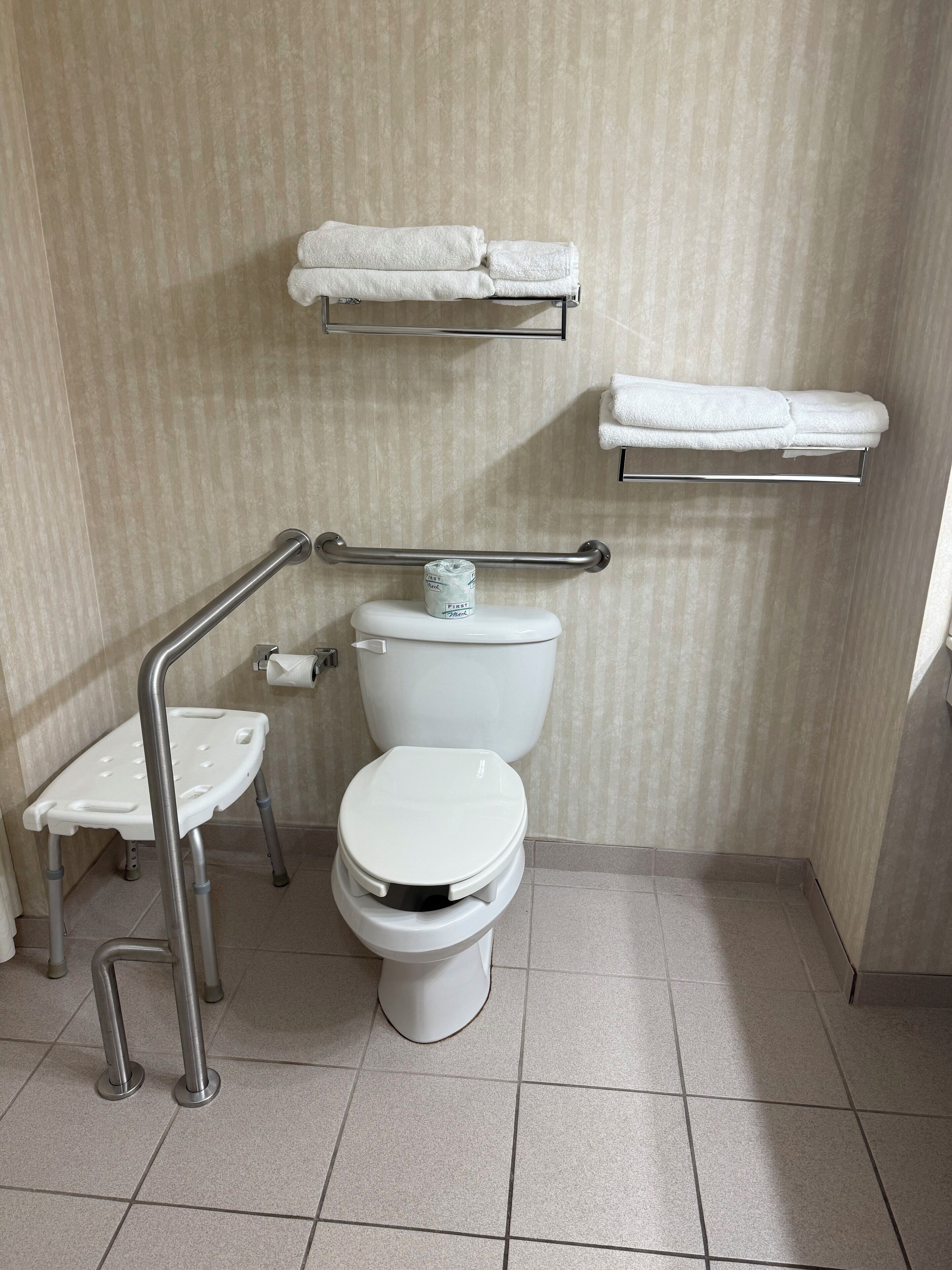 Une salle de bain équipée de toilettes, d’un lavabo et d’une barre d’appui. Deux porte-serviettes sont fixés au mur.