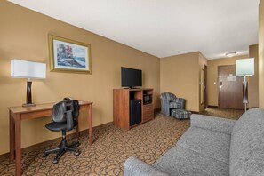 Habitación de hotel con escritorio, silla de oficina, sofá, televisión y una pequeña kitchenette.