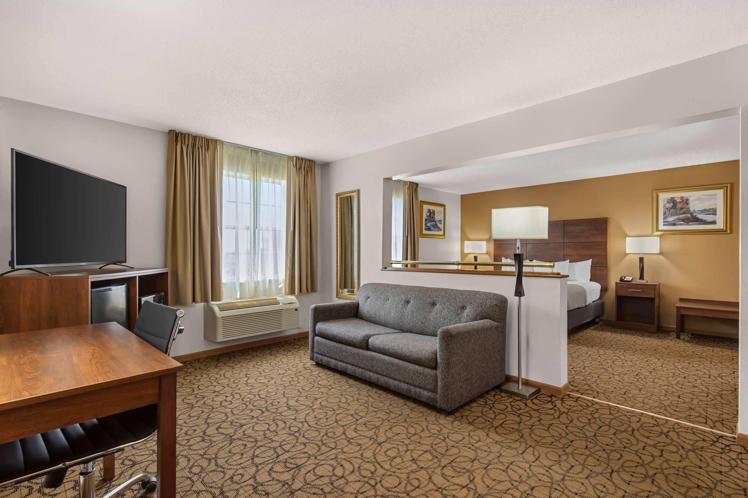 A hotel room with a flat-screen TV, a sofa, a desk with a chair, a bed, a bedside table with a lamp, and a patterned carpet.